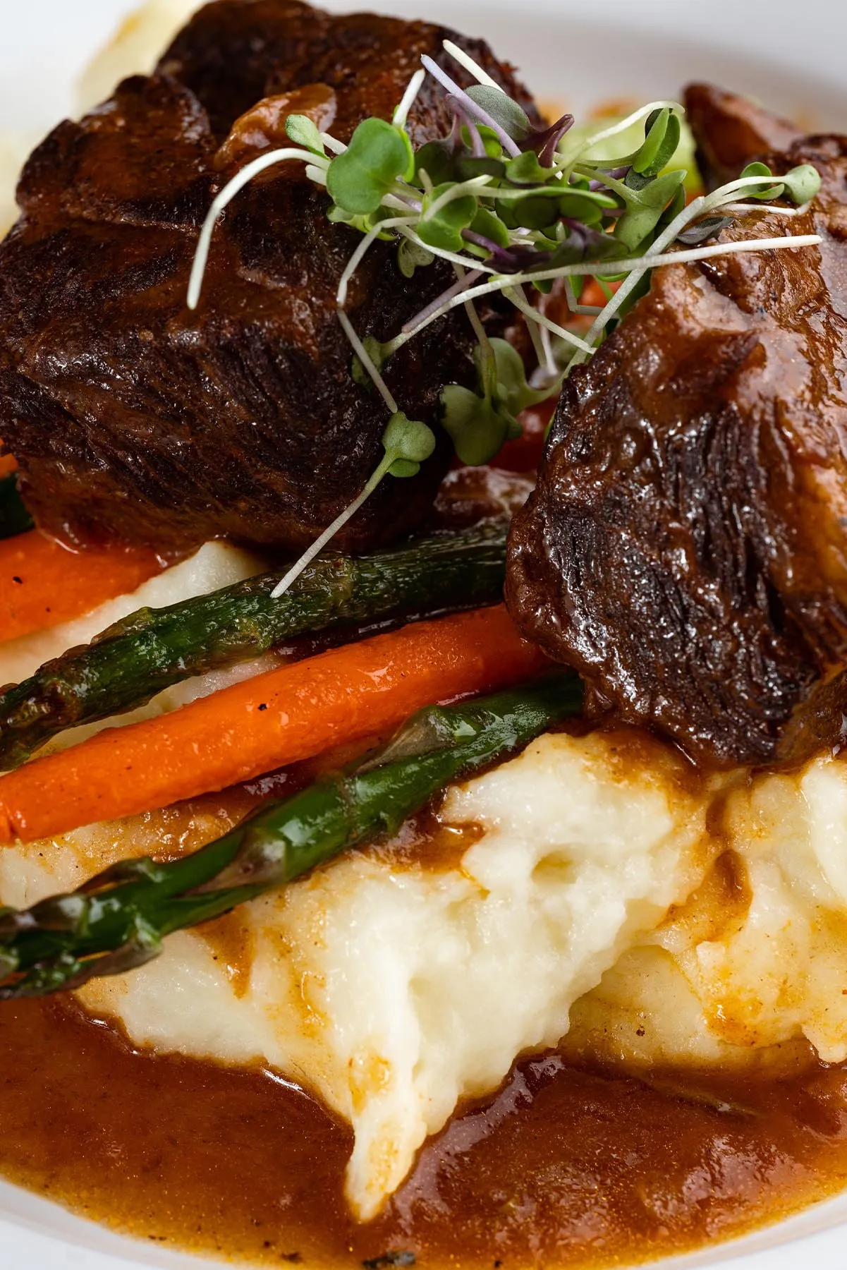 Braised beef short ribs atop creamy mashed potatoes with carrots, asparagus, and microgreens garnish in brown sauce.