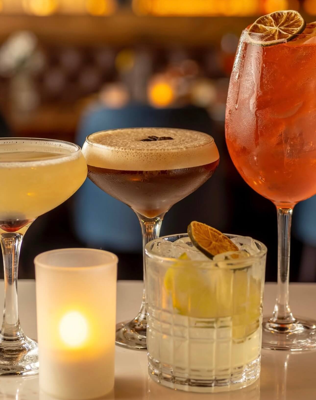 Four assorted cocktails on a table with a lit candle in a frosted holder in the foreground.